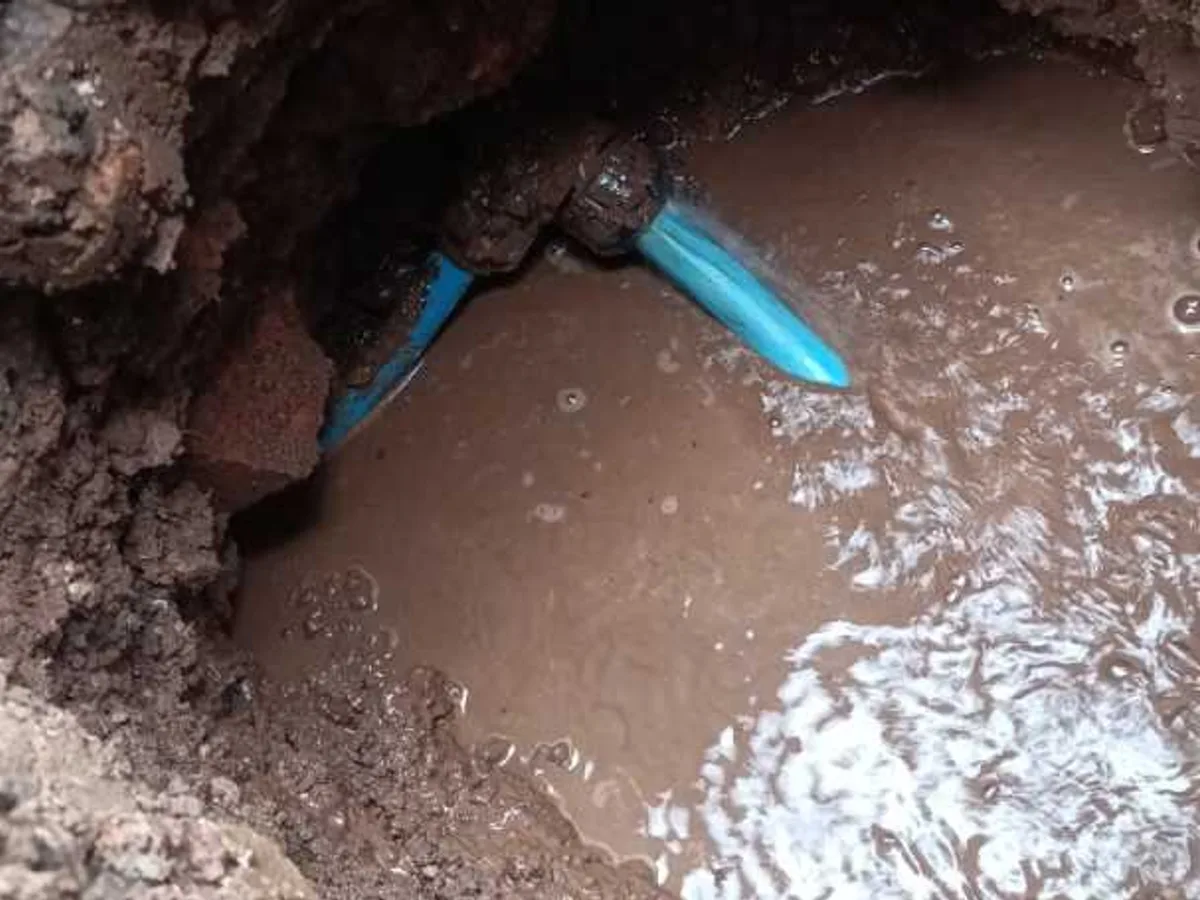 A blue plastic supply pipe leaking into a flooded excavation, water still escaping from the joint.